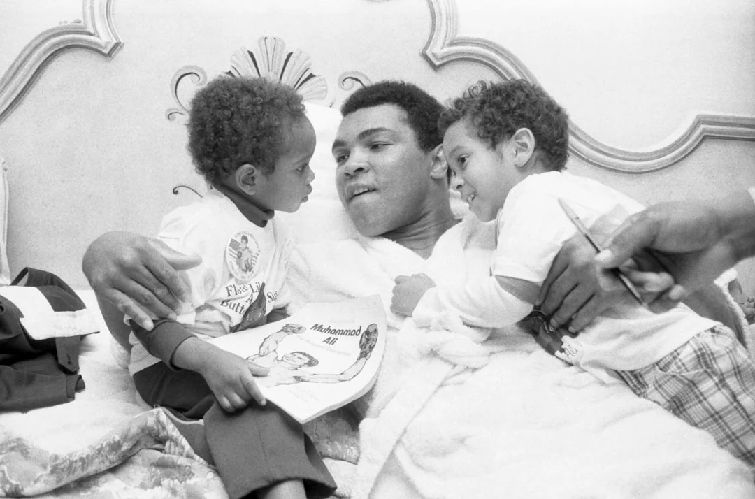 A man in a robe lies in bed with two young children. One child is holding a book titled "Muhammad Ali." They are all smiling and engaged in conversation, creating a warm, familial scene.