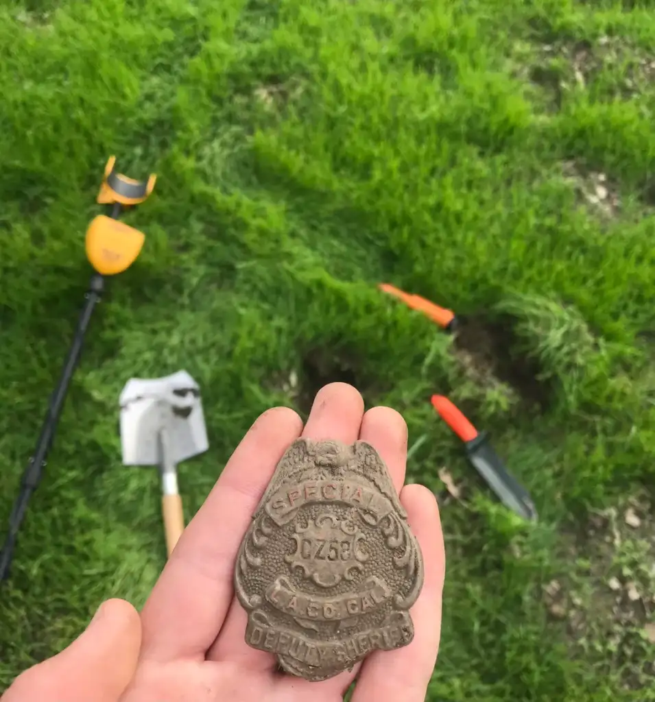 A person holds an old badge labeled "Special Deputy Sheriff" in their hand. In the background, a metal detector, a shovel, and a digging tool are on grassy ground, with a small hole nearby.