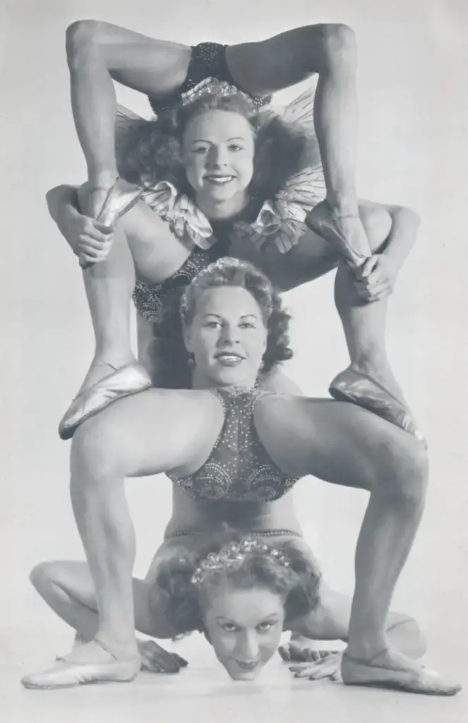 Three female acrobats balance in a vertical formation. The top athlete smiles while performing a flexible pose, the middle performer holds a leg-splitting position, and the bottom supports her teammates, all dressed in sparkling leotards.