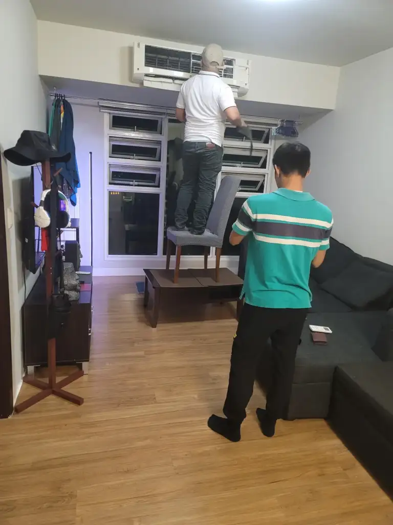 Two people in a room are working on an air conditioning unit. One person stands on a chair and table, reaching up to the unit, while the other stands nearby holding tools. The room has a couch, coat rack, and wooden floor.