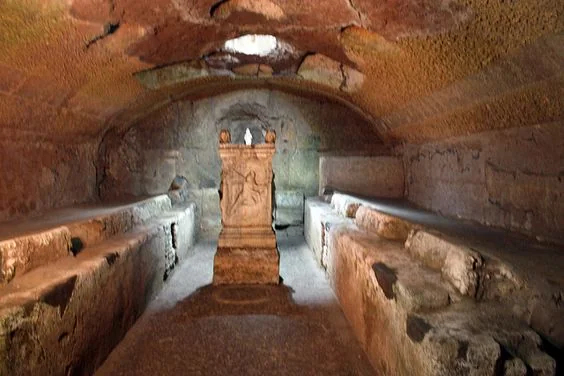 Ancient stone chamber with an arched ceiling, featuring worn benches along the sides and a detailed stone altar at the center. The scene is dimly lit, highlighting the weathered textures and architectural features.