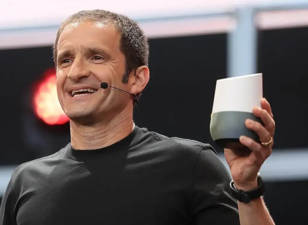 A person wearing a black shirt is holding a smart speaker while speaking into a headset microphone, with a red light in the background.