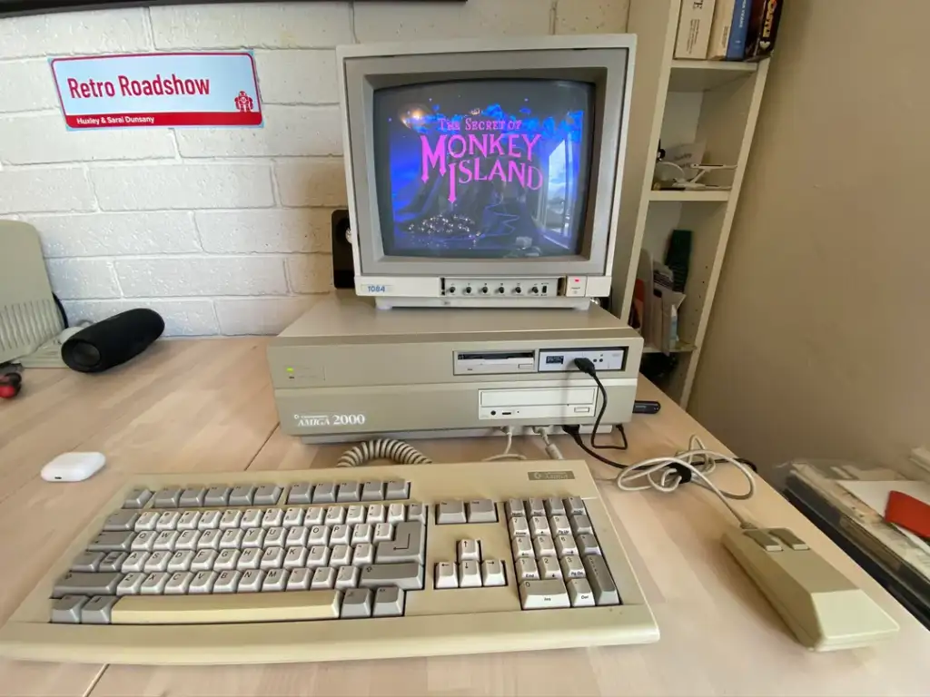 A vintage computer setup featuring an Amiga 2000 with a keyboard and mouse on a wooden desk. The monitor displays the title screen of "The Secret of Monkey Island." A retro roadshow sign is mounted on a white brick wall.