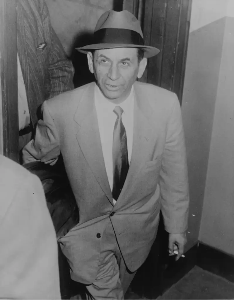 A man wearing a suit, tie, and fedora hat walks through a doorway. He appears to be in an indoor setting, possibly an office or hallway. The photo has a vintage, black-and-white appearance.