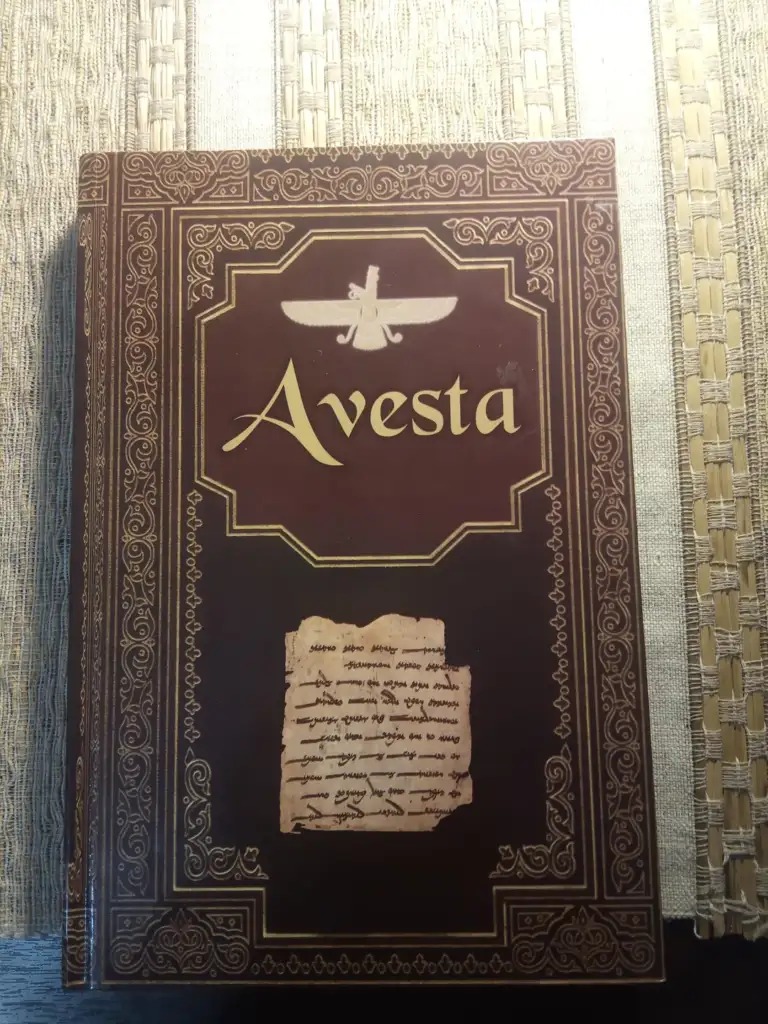 A decorative book cover titled "Avesta" with intricate gold patterns and a small image of ancient script. The design features Zoroastrian symbols and motifs on a brown background, placed on a woven surface.