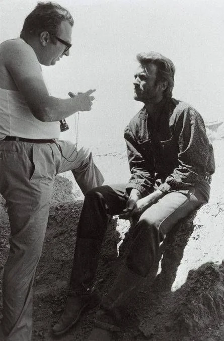 Two men on a movie set discuss; one in a tank top gestures while standing, the other sits on a rock wearing a denim shirt and boots, listening. It's a sunny day with a barren, rocky backdrop. Black and white photo.