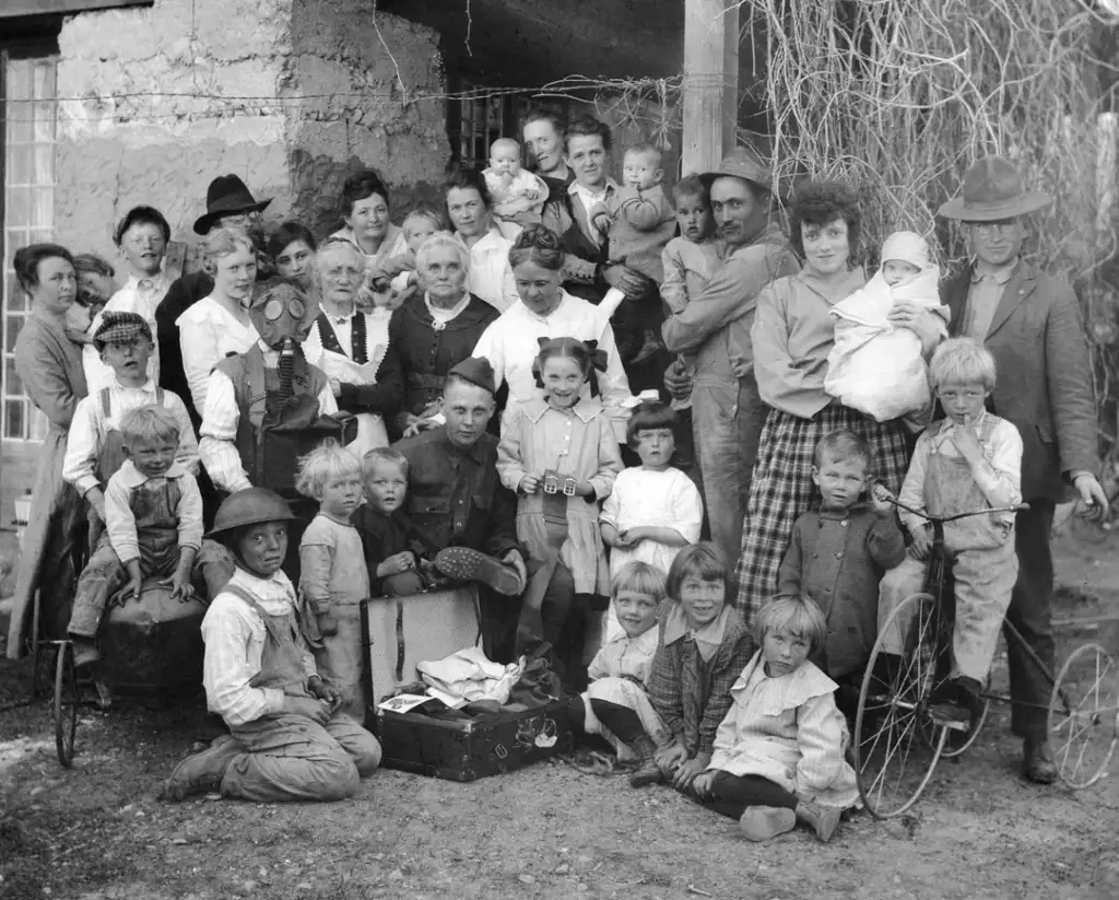 A large vintage black and white photo of a group of people, including men, women, and numerous children, gathered outdoors. Some children have bicycles, and there is an open suitcase with items. The group appears to be posed for the photograph.