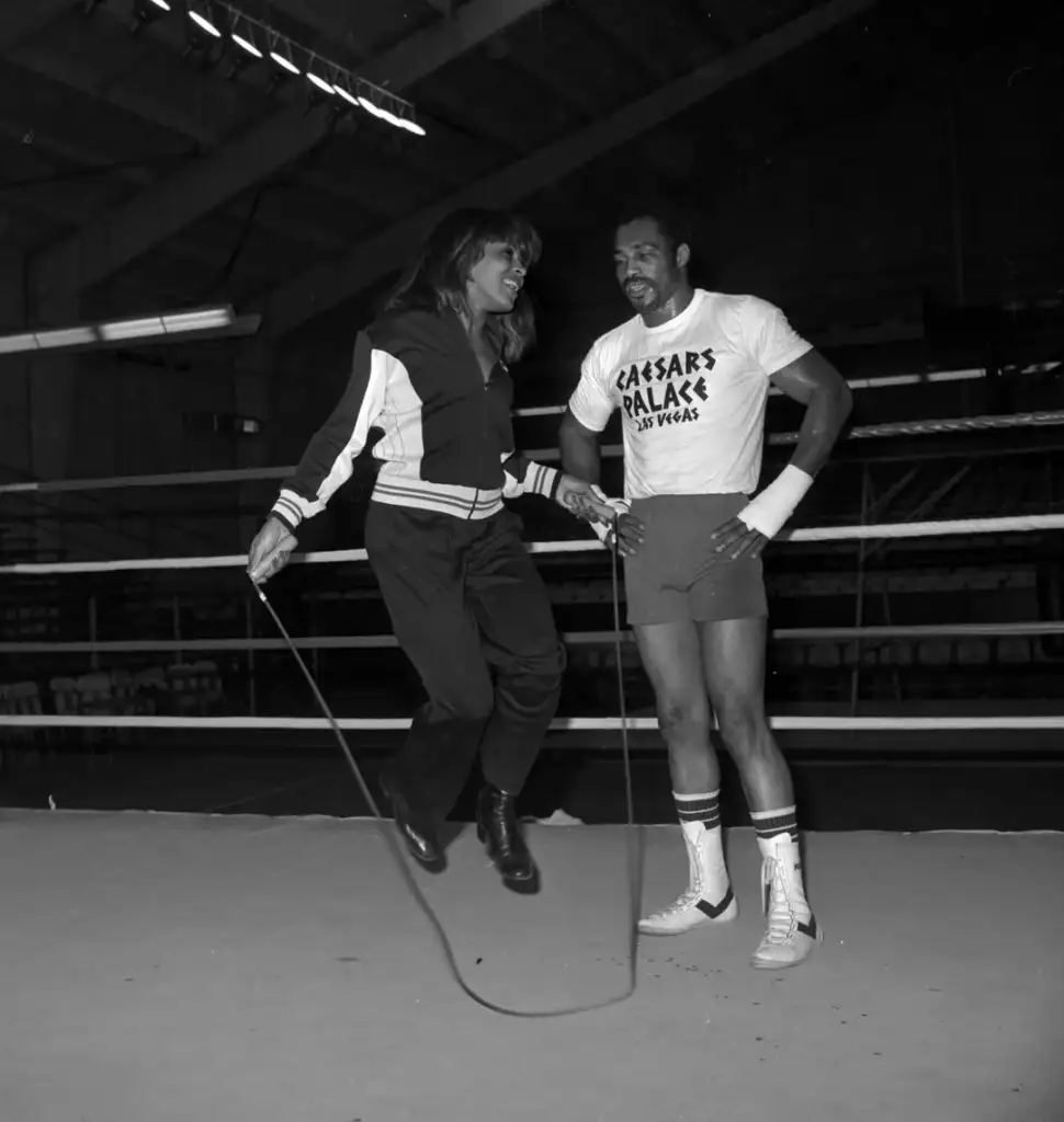 A woman jumps rope in a boxing ring wearing a tracksuit. Beside her, a man in boxing gear, with a Caesar's Palace T-shirt, watches and smiles. The background shows ropes and audience seating, suggesting a gym or training environment.