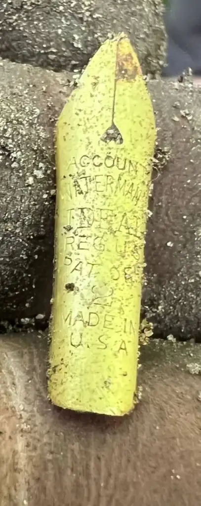 A close-up of a worn yellow pen nib with visible dirt. The nib has engraved text: "Account Waterman Ideal Reg U.S. Pat Off Made in U.S.A." It's held between two fingers, with a background of a textured surface.