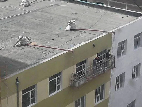 Two workers stand on a suspended platform on the side of a building. The platform is secured to the roof by large sandbags, creating an unusual safety setup. They appear to be working on the building's exterior wall.