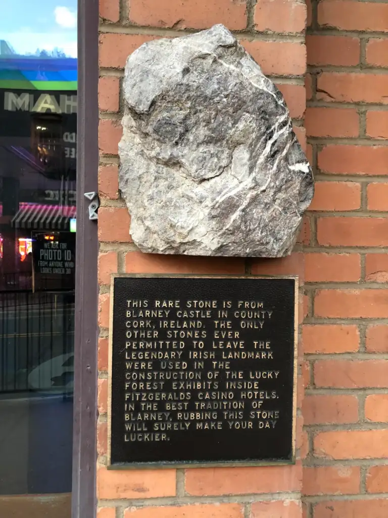 A piece of stone is mounted on a brick wall above a plaque. The plaque explains that the stone is from Blarney Castle in Ireland and was used in the construction of casino hotels in Fitzgeralds, promoting luck when rubbed.