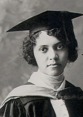 Black and white photo of a young woman in a graduation cap and gown. She has short curly hair and is looking directly at the camera with a neutral expression. The background is a simple studio backdrop.