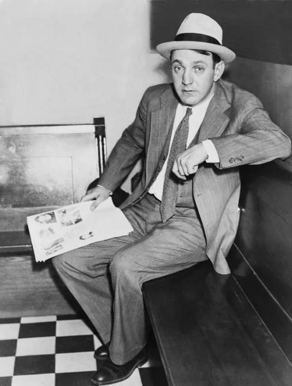 A man in a suit and tie sits on a wooden bench, holding a newspaper. He wears a light-colored fedora and looks towards the camera. The floor has a checkered pattern, and there is a wooden panel behind the bench.