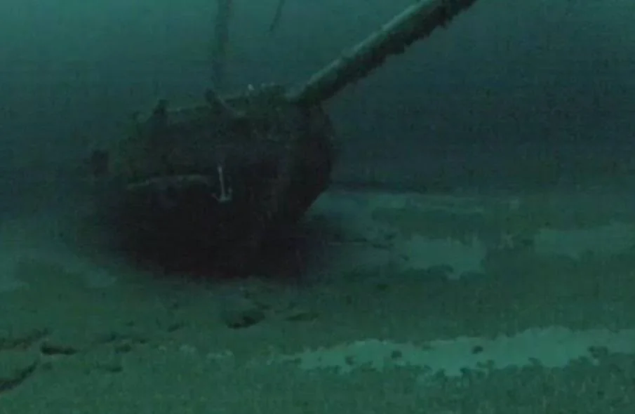 Underwater image of a shipwreck resting on a sandy seabed. The vessel is partially buried, with some parts covered in sediment. The water around the shipwreck is murky, giving an eerie and mysterious atmosphere.