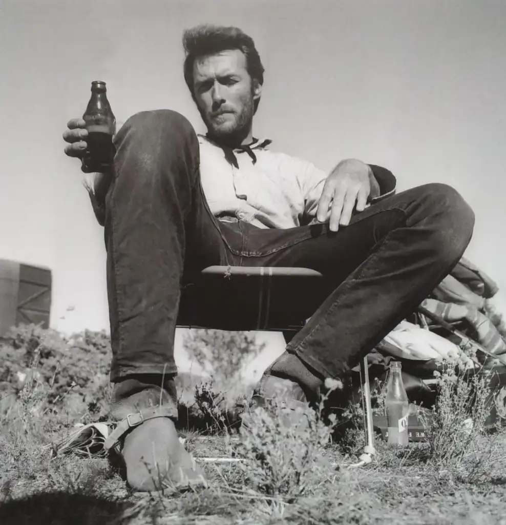 A man sitting on a chair outdoors, holding up a bottle in one hand. He wears a casual shirt and jeans, with one leg crossed over the other. Sparse vegetation and a clear sky are visible in the background. The image is in black and white.