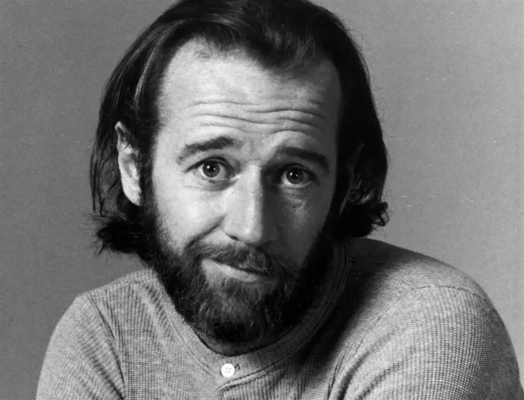 Black and white portrait of a man with medium-length hair and a beard, looking directly at the camera with a slight smile. He is wearing a buttoned shirt. The background is plain.