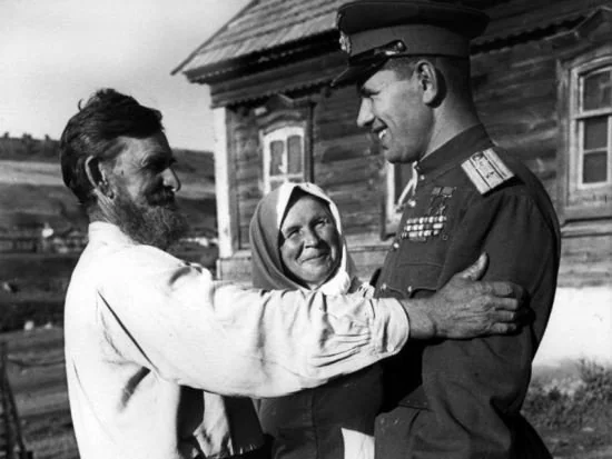 An older man and woman warmly embrace a man in a military uniform covered in medals. They stand outside a wooden house on a sunny day. The military man smiles, looking at the woman, who is wearing a headscarf.