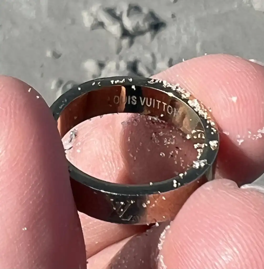 Close-up of a hand holding a gold ring with "Louis Vuitton" engraved on the inside. The hand is covered with sand, and the background is a sandy surface.