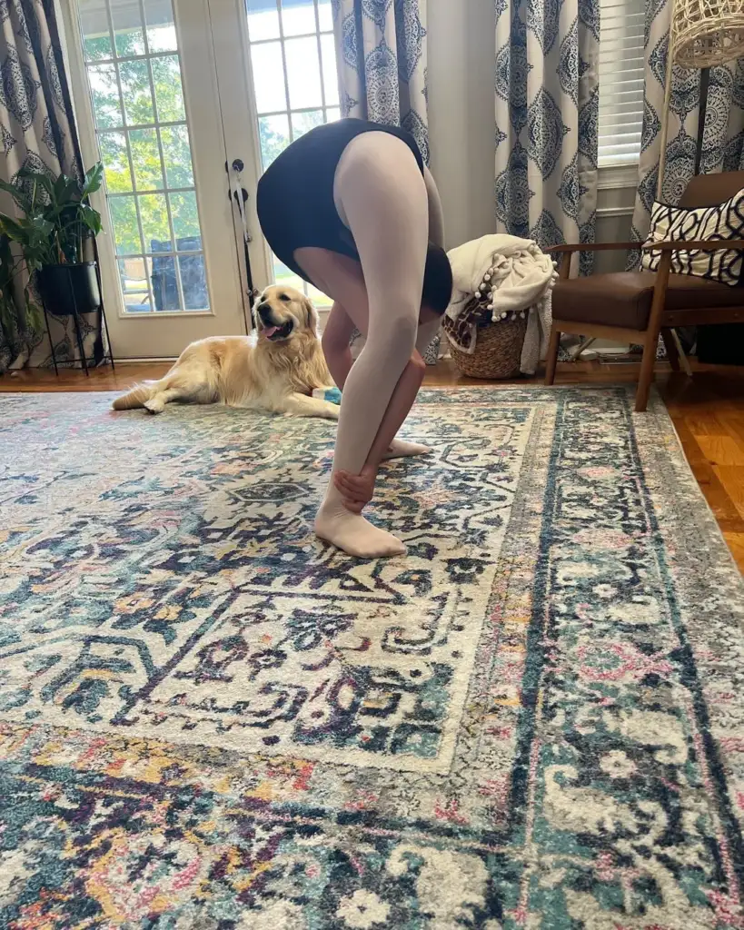 A person wearing a black leotard and white tights bends over in a room with a patterned rug. A golden retriever lies on the floor near a large window with curtains, looking in their direction.