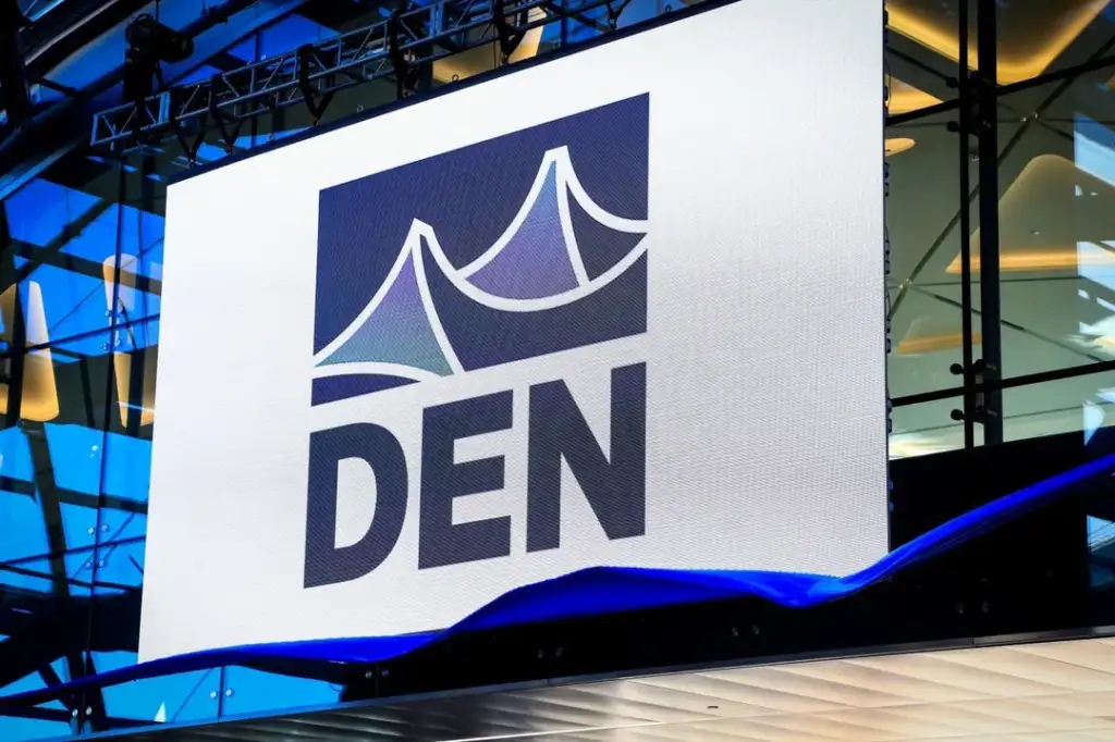 A large digital screen displays the logo and letters "DEN" in a modern architectural setting with glass surfaces and reflections. Blue light highlights elements of the scene, and ceiling structures are visible in the background.