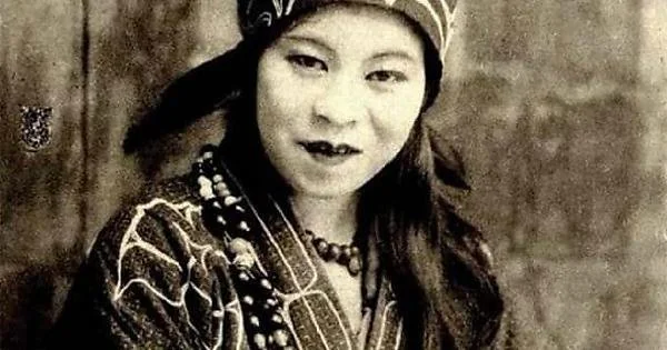 A black-and-white photo of a person wearing a patterned headband and traditional clothing adorned with beaded necklaces. The expression is calm, with a subtle smile. The background is blurred.