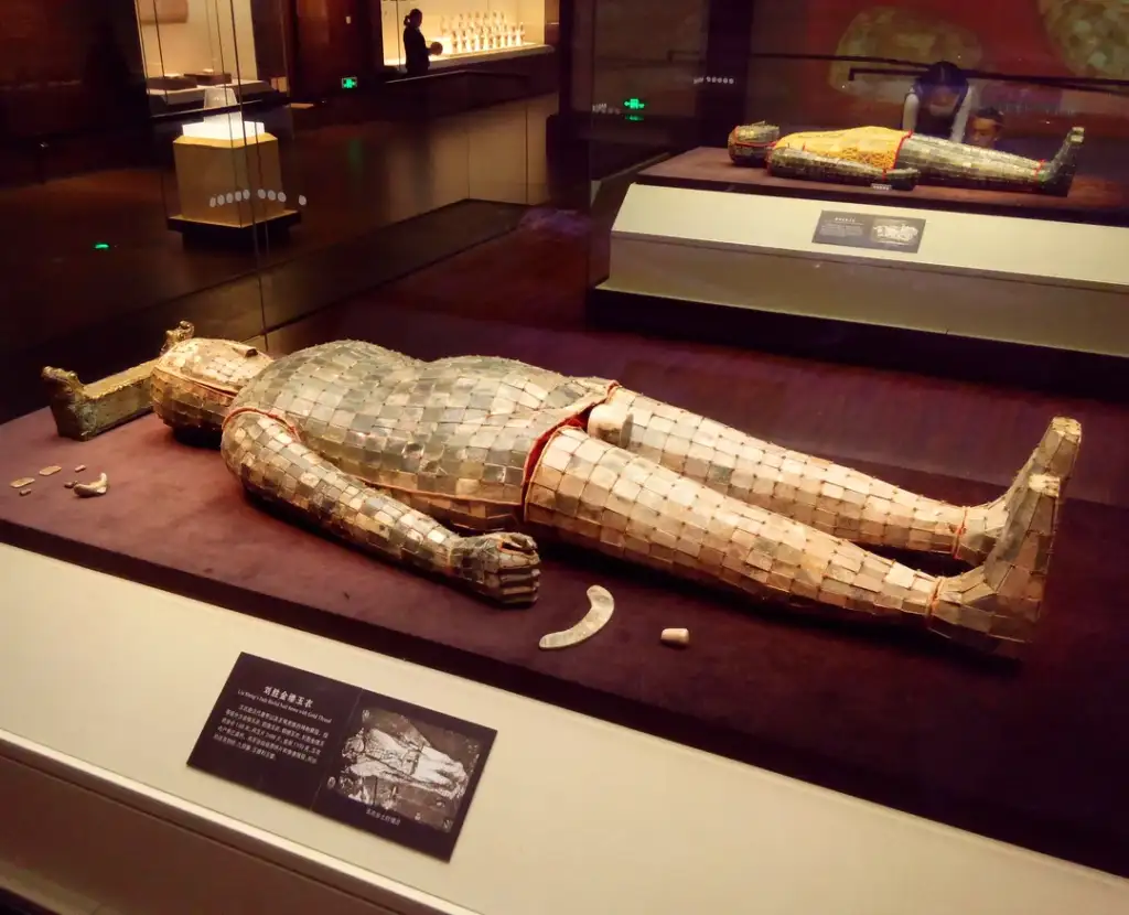 A museum exhibit displaying an ancient burial suit made of jade tiles sewn together with red thread, laid flat in a glass case. Another similar suit is visible in a distant case. Informational plaques are on the display.