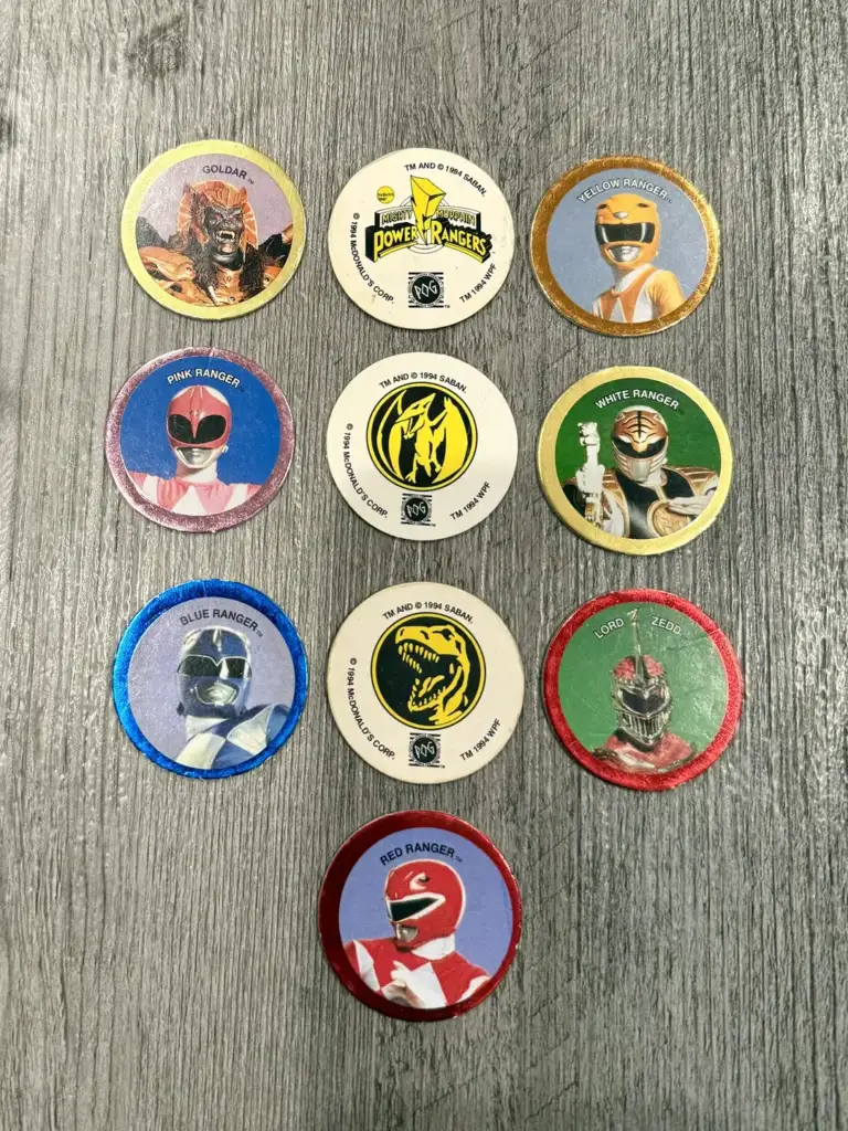 Nine Power Rangers-themed pogs are arranged on a wooden surface. Each pog features different characters and logos, showcasing various Power Rangers and a villain.