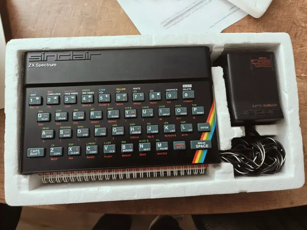 The image shows a ZX Spectrum computer in its original packaging. The black keyboard features colored keys with various commands. A power adapter and cables are also included. The packaging is white foam, and a piece of paper is partially visible.