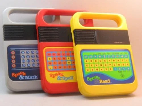 Three vintage educational electronic toys are shown: grey "Speak & Math," red "Speak & Spell," and yellow "Speak & Read." Each has a handle and colorful buttons on the front, featuring letters and symbols.