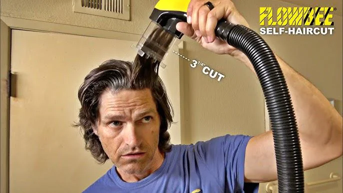 Man using a Flowbee self-haircut device on his long, wavy hair. The vacuum-like attachment is trimming his hair with a labeled "3/4 inch cut" guide. He is wearing a blue shirt and standing in a room with a beige wall.