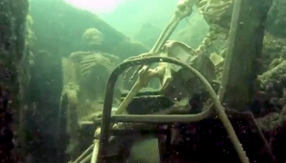 Underwater scene showing two artificial skeletons sitting in lawn chairs, one with its hand on an object resembling a steering wheel. Surroundings are murky with greenish light, giving an eerie aspect to the scene.