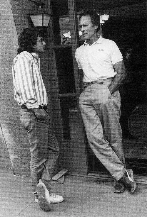 Two men are standing and talking outside a building. One is wearing a striped shirt and jeans; the other is in a polo shirt and slacks. Both have their hands in their pockets, and one man's foot is resting on the wall behind him.