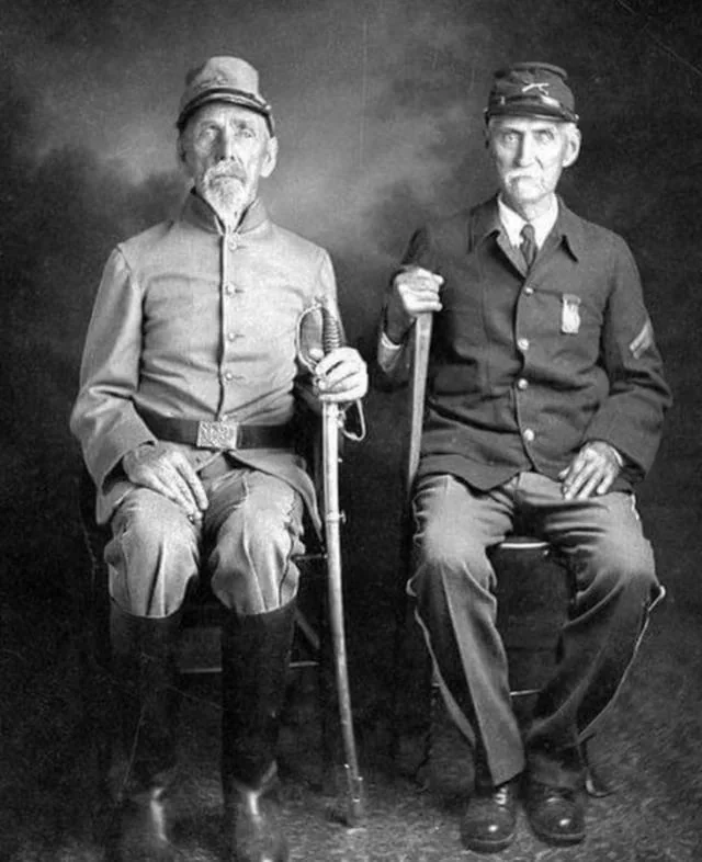 Two elderly men in historical military uniforms sit side by side. The man on the left wears a Confederate uniform and holds a sword. The man on the right wears a Union uniform and holds a cane. Both have white beards and appear serious.