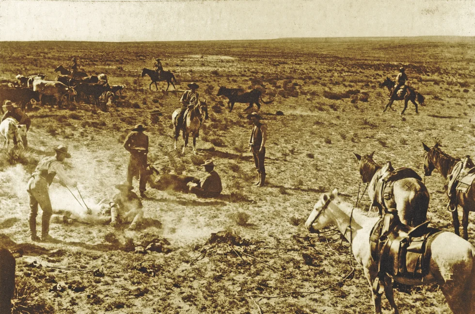 A sepia-toned image of cowboys herding cattle on a vast, open plain. Several cowboys on horseback surround a group branding a cow on the ground. Horses are scattered across the rugged terrain under a wide, barren sky.