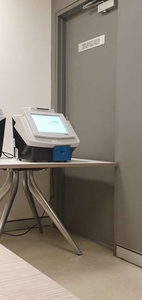 A digital voting machine positioned on a wooden table beside a gray door with a sign saying "Fire Safety Door Do Not Block." The area has a plain, professional setup with neutral-colored walls and floor.