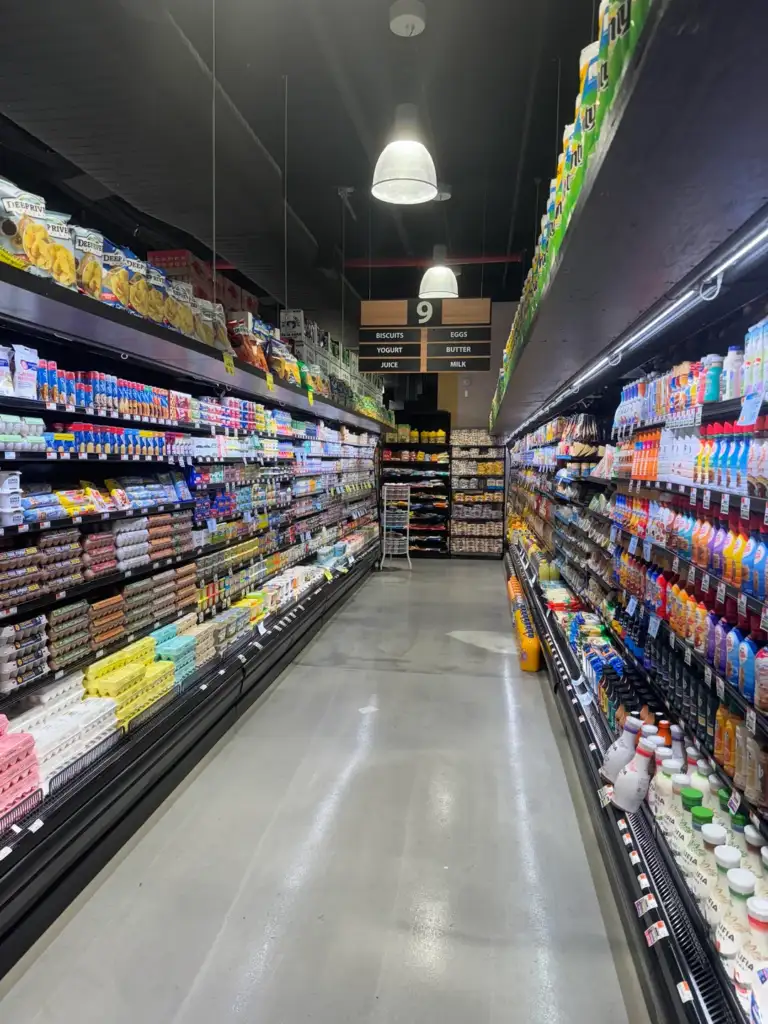 A grocery store aisle filled with various products. Shelves on both sides are stacked with eggs, milk, and other packaged goods. Overhead lights illuminate the aisle, and a sign above indicates this is aisle 9, featuring bread and dairy items.