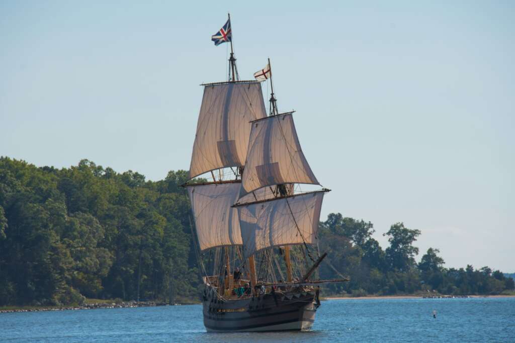 A historic-style sailing ship with multiple masts and sails unfurled, flying a Union Jack flag, glides on a calm body of water. Lush green trees line the shore under a clear blue sky.