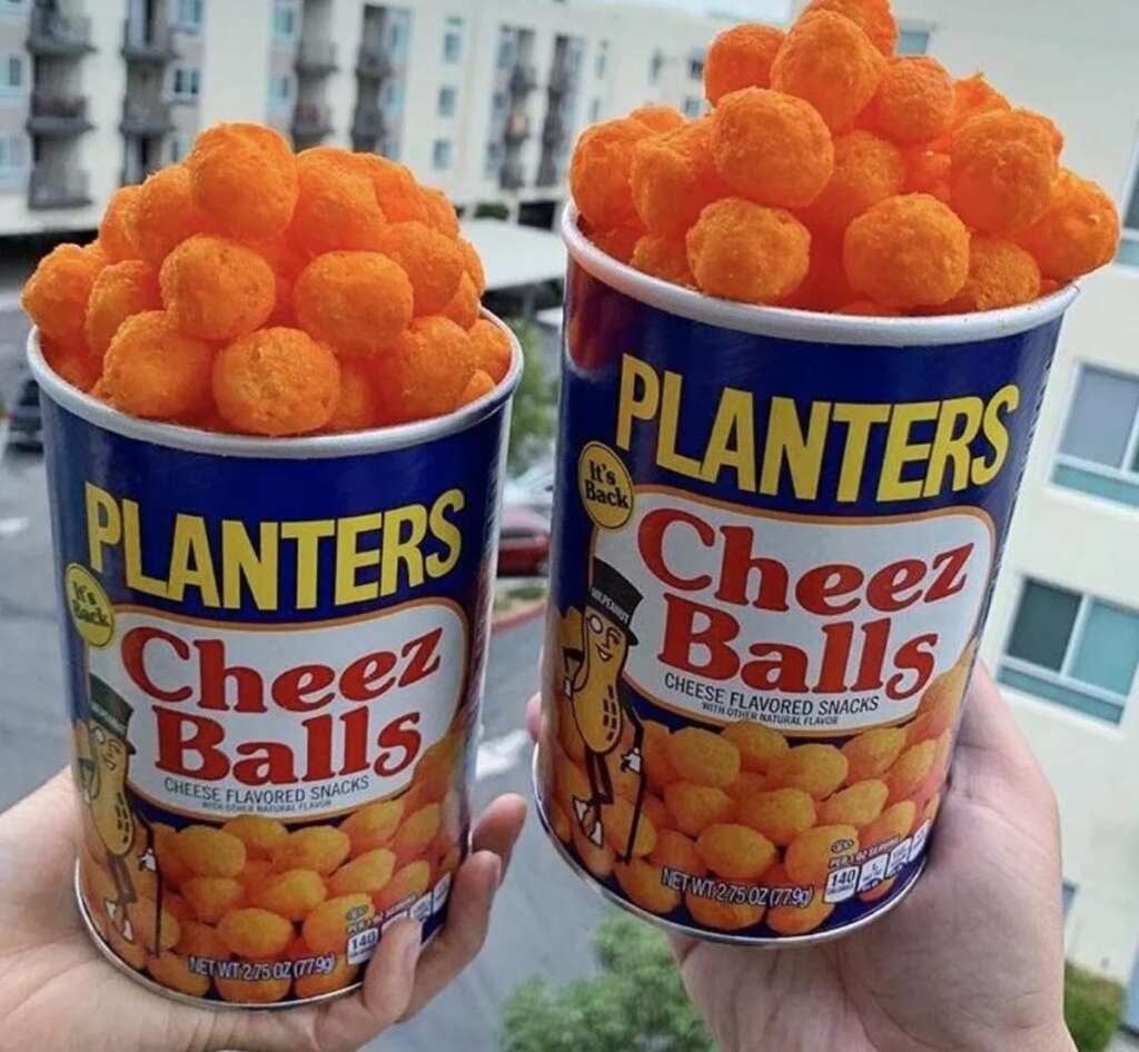 Two hands holding cans of Planters Cheez Balls in front of a blurred urban building background. Each can is overflowing with bright orange cheese balls. The label features a cartoon character with a monocle and top hat.