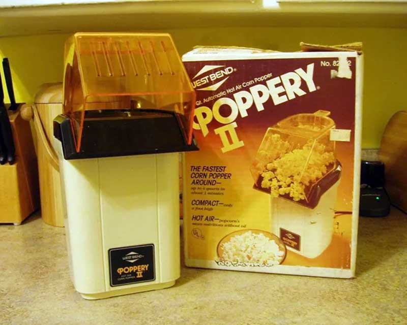 A vintage West Bend Poppery II hot air popcorn popper sits on a kitchen counter next to its original box. The popper has a white base and transparent orange lid. The box features an image of popcorn overflowing from the machine.