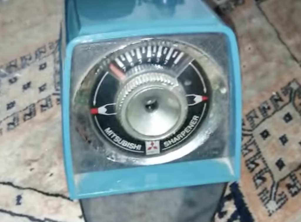 A close-up of a vintage Mitsubishi pencil sharpener, showing the dial and blade mechanism. The metallic details stand out against the blue casing, and it rests on a patterned rug.