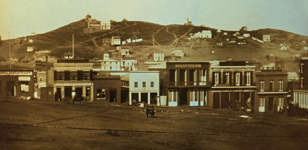 A historical photograph of a small town with a row of wooden buildings in front of a hill. The storefronts include signs for various businesses. Sparse structures are scattered on the hill in the background, and a few people and horses are visible.