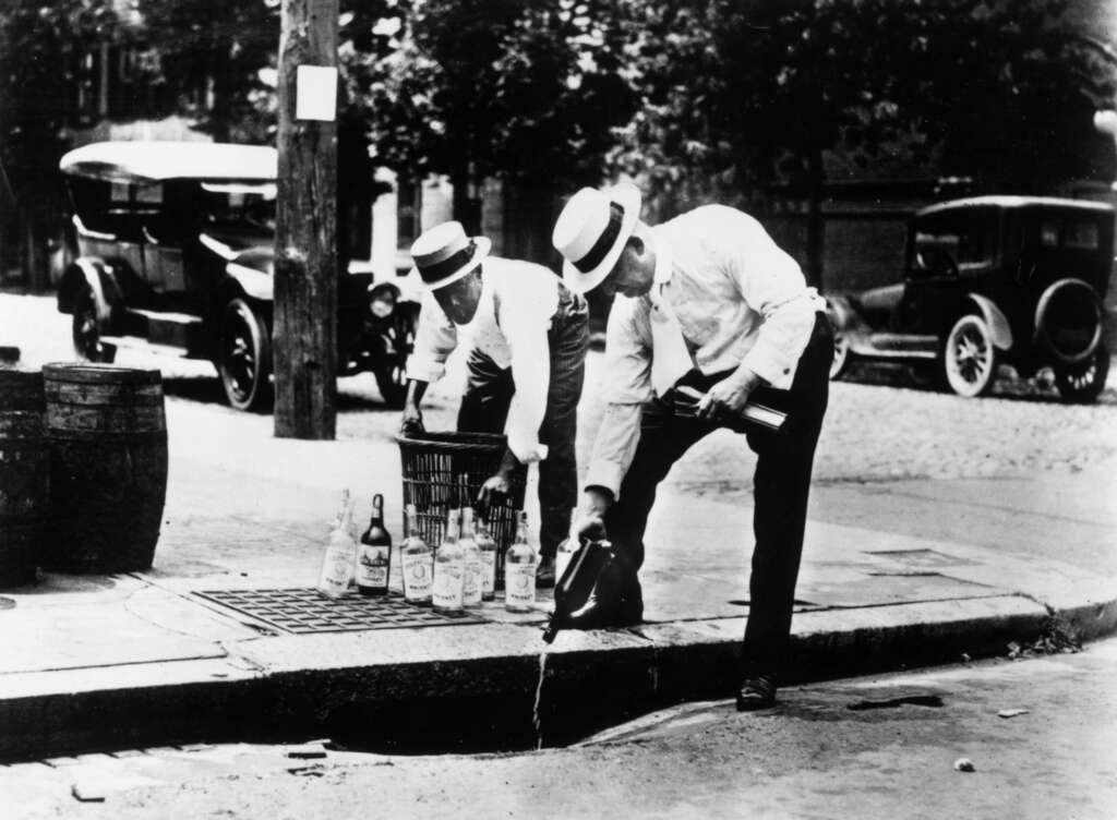 Two men in white shirts and hats pour bottles of alcohol into a street drain during Prohibition. They stand on a sidewalk with vintage cars and trees in the background.