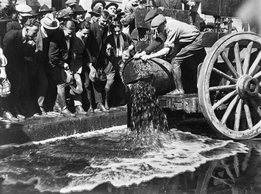 A historical black-and-white photo showing a crowd of people watching as a worker empties a barrel of liquid, likely alcohol, from a cart onto the street. The scene suggests prohibition-era confiscation or disposal.