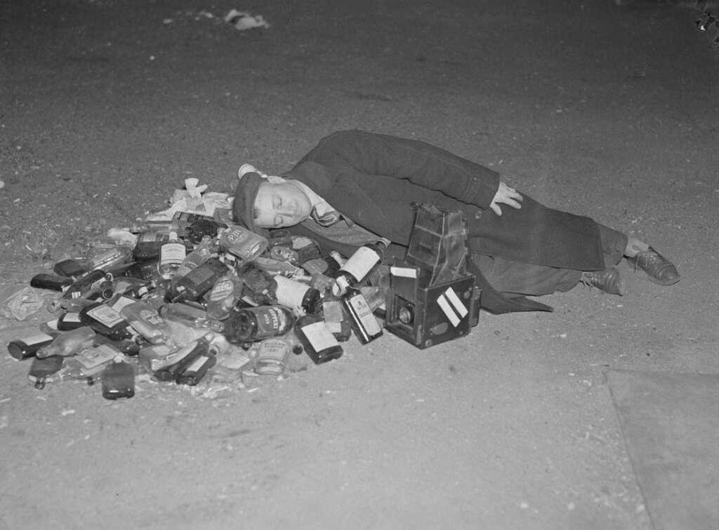 A man in a coat and hat lies asleep on the ground next to a pile of empty alcohol bottles and cartons. The scene appears to be outside, at night.