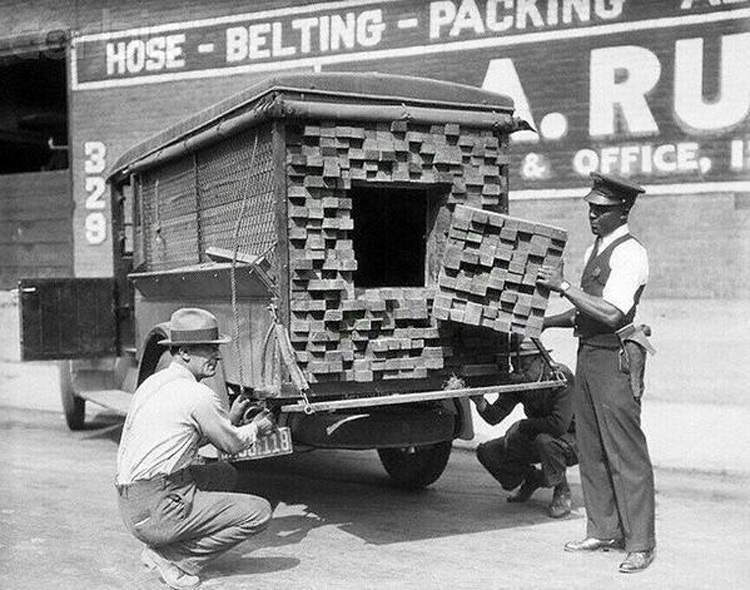 Two men unload bricks of grass from the back of a vintage delivery truck. A large rectangular opening is visible in the stacked grass bricks. Another person is partially visible underneath the truck. A building with signs is in the background.