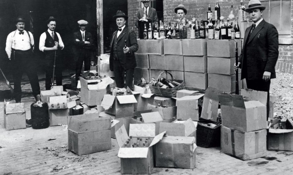 A black and white photo shows several men in early 20th-century attire standing around open crates and boxes filled with bottles of alcohol. The scene appears to be from the Prohibition era, with boxes stacked against a brick wall in the background.