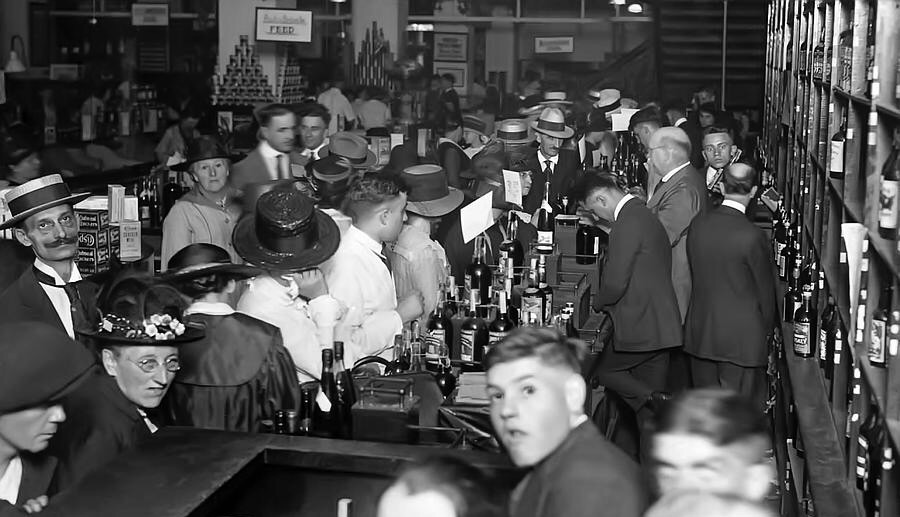 Black and white photo of a crowded 1920s bar. Men and women in period attire, including hats and suits, gather around the bar counter filled with bottles. The lively scene suggests a bustling atmosphere typical of the Prohibition era.