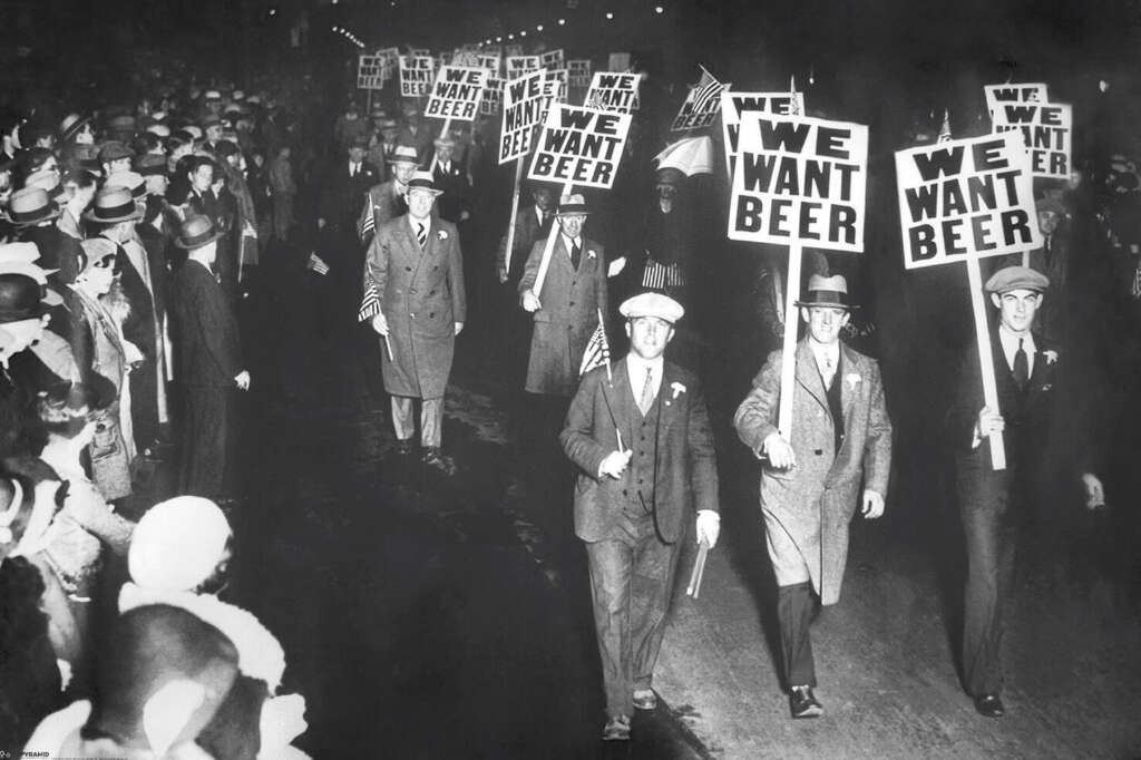 A black-and-white photo shows a group of men in coats and hats marching on a street at night, holding signs that read "We Want Beer." A large crowd is gathered along the sides, watching the march.