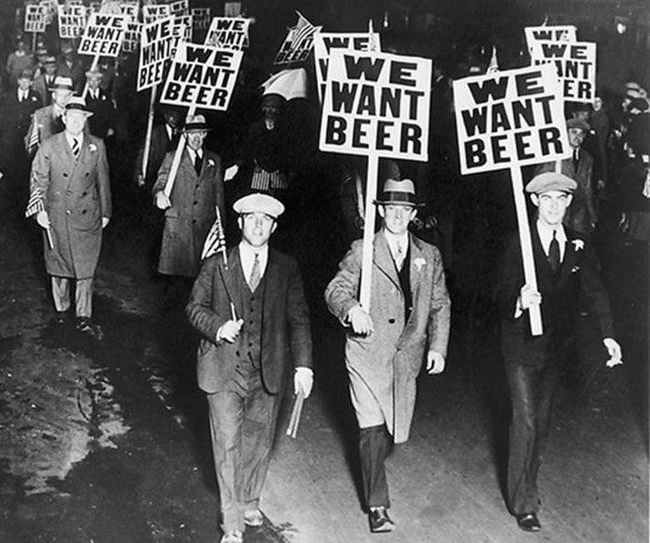 A historical black-and-white photo of a crowd of men marching at night, holding signs that read "WE WANT BEER." The men are dressed in suits and hats, and the protest reflects the Prohibition era.