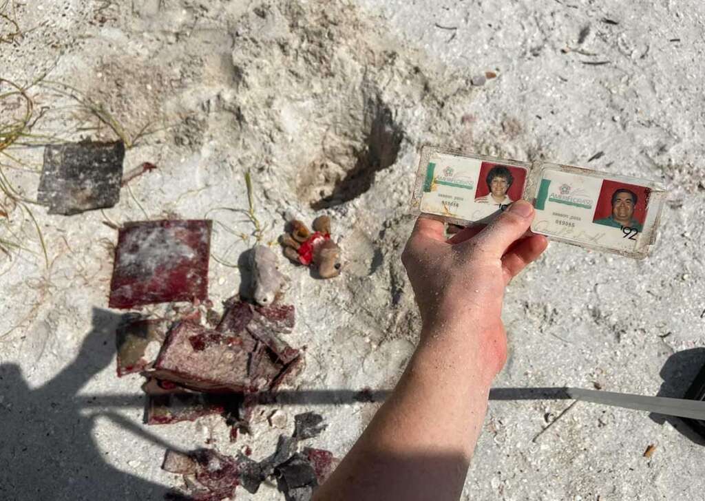 A hand holds two identification cards above a sandy ground. Nearby, there are scattered burned documents and a small toy bear.